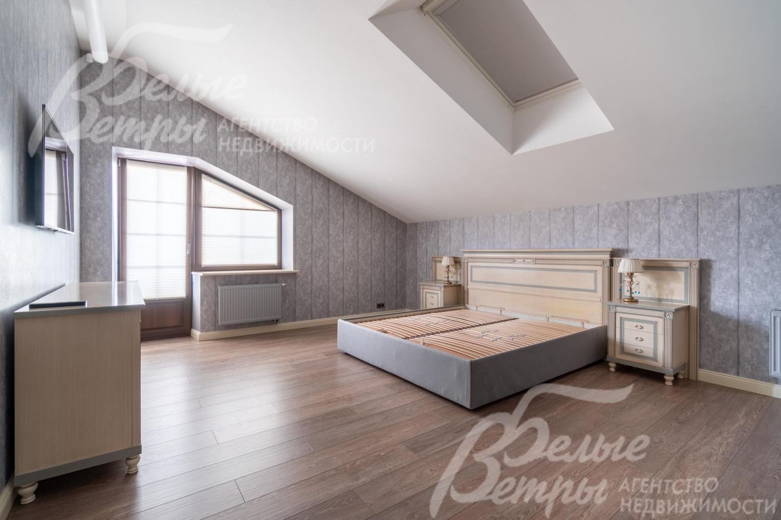 Просторная спальня на втором этаже с мансардной крышей и скошенными окнами оформлена в спокойных серо-бежевых тонах: интерьер в классическом стиле, доска из дуба на полу добавляет уюта